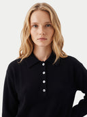 A blonde woman wearing a black long-sleeve knit polo shirt with white buttons, photographed against a clean light gray studio background.