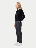 A woman stands in profile wearing a black long-sleeve henley top, dark charcoal gray trousers, and white sneakers against a light gray studio background.