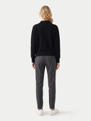 A woman viewed from the back wearing a black long-sleeve mock-neck top, dark gray trousers, and white sneakers, standing against a light gray studio background.