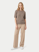 A woman modeling a short-sleeve ribbed knit sweater in taupe brown, paired with beige wide-leg trousers and white sneakers, against a light gray studio background.