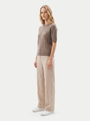 A woman stands in profile wearing a short-sleeve ribbed knit sweater in taupe-brown, wide-leg beige trousers, and white sneakers against a clean light gray studio background.