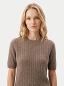 A blonde woman wearing a short-sleeve crewneck knit sweater in a warm taupe-brown color, shown against a plain light gray studio background.