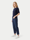 A woman modeling a dark navy ribbed short-sleeve top and dark blue jeans with white sneakers, standing against a light gray studio background.