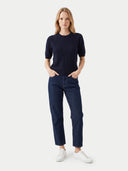 A woman modeling a dark navy ribbed short-sleeve knit top and dark blue jeans with white sneakers, standing against a light gray studio background.