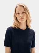 A blonde woman wearing a dark navy blue short-sleeve cable-knit sweater, photographed against a clean light gray studio background.