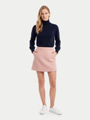 A woman modeling a navy blue turtleneck sweater and a light pink mini skirt with white sneakers, standing against a light gray studio background.