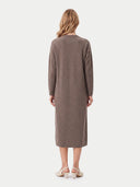 A woman viewed from the back wearing a long-sleeve, knee-length taupe knit dress and light-colored heeled sandals against a plain light gray studio background.