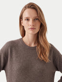 A woman with long light brown hair wearing a dark brown crewneck knit sweater, photographed against a clean off-white studio background.