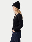 A woman in profile wearing a black ribbed knit beanie, a black long-sleeve V-neck sweater, and dark blue jeans, photographed against a light gray studio background.