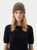 A woman wearing a brown ribbed knit beanie with a green embroidered clover logo, paired with a black ribbed short-sleeve top, against a light gray background.
