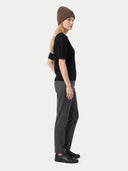 side view of a woman standing wearing a olive brown beanie with brand logo, a black short sleeve top, dark gray pants, black sneakers on a light gray background.