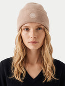 A woman with blonde hair wearing a ribbed beige beanie with a small white embroidered logo, paired with a black V-neck sweater, against a light gray background.