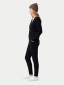 A woman in a side profile wearing a brown knit beanie, a black long-sleeve top, black pants, and white sneakers, standing against a light gray studio background.