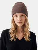 A woman with blonde hair wearing a ribbed brown beanie with an embroidered logo and a black V-neck sweater, photographed against a light gray background.