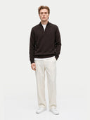 front view of a man standing wearing a dark brown quarter zip cardigan over a white t-shirt and white pants on a light gray background.