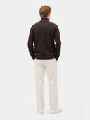 A man viewed from behind wearing a dark brown mock-neck knit sweater, off-white trousers, and white sneakers, standing against a plain light gray studio background.
