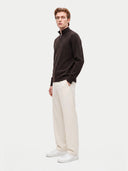 side view of a man standing wearing a dark brown quarter zip cardigan over a white t-shirt and white pants on a light gray background.