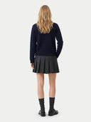 A woman viewed from behind wearing a dark navy cable-knit sweater, a dark gray pleated mini skirt, dark gray socks, and black leather shoes, against a light gray studio background.