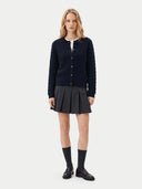 A model stands against a light gray studio background wearing a dark navy textured cardigan over a white collared shirt, paired with a dark gray pleated mini skirt, black knee-high socks, and black leather shoes.