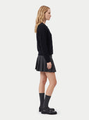 A woman stands in profile wearing a black textured long-sleeve top, a black pleated mini skirt, black knee-high socks, and black leather loafers against a light gray studio background.