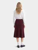 A woman shown from the back wearing a white long-sleeve button-up shirt and a knee-length dark burgundy skirt, paired with white socks and dark loafers, against a light gray studio background.