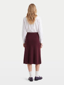 A woman shown from the back wearing a white long-sleeve button-up shirt and a knee-length burgundy skirt, paired with white socks and dark loafers, against a plain light gray studio background.