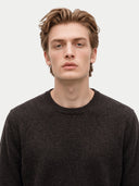 A close-up portrait of a young man with light brown hair wearing a dark charcoal gray knit crewneck sweater, set against a plain light gray background.