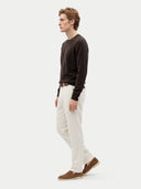 A man in profile wearing a dark brown long-sleeve knit sweater, off-white trousers, a brown belt, and brown suede shoes, standing against a light gray studio background.