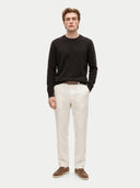 A man modeling a dark brown long-sleeve knit sweater, off-white linen trousers, a brown belt, and brown suede shoes against a light gray studio background.