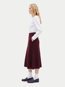 A woman stands in profile wearing a white long-sleeve top, a dark burgundy midi skirt with side pockets, white socks, and dark purple loafers, against a light gray studio background.