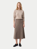 A woman stands against a light gray studio background wearing a beige crewneck sweater, a taupe-brown midi skirt, and black leather loafers.