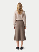 A woman viewed from the back wearing a light beige knit sweater and a brown midi skirt, paired with black loafers, against a plain light gray studio background.