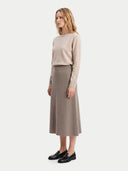 A woman stands in profile wearing a light beige long-sleeve knit sweater and a taupe midi skirt, paired with black loafers, against a plain light gray studio background.
