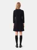 A woman viewed from the back wearing a black long-sleeve mini dress, black knee-high socks, and black shoes against a light gray studio background.