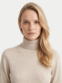A blonde woman wearing a light beige ribbed turtleneck sweater, photographed against a clean off-white background.