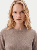 A close-up shot of a woman with blonde hair wearing a light brown crewneck knit sweater against a plain light gray background.
