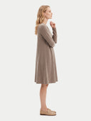 A woman stands in profile wearing a long-sleeve, knee-length knit dress in a taupe-brown color, paired with beige loafers, against a solid off-white background.