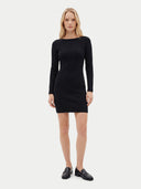 A blonde model stands against a light gray studio background wearing a fitted black long-sleeve mini dress with a boat neckline and black leather loafers.