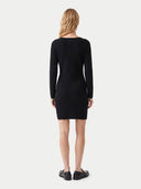 A woman viewed from the back wearing a fitted black long-sleeve mini dress and black loafers against a plain light gray studio background.