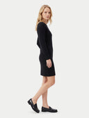 A woman modeling a black long-sleeve knit mini dress, shown in profile against a light gray studio background. She is wearing black leather loafers.