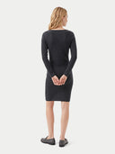 A woman viewed from the back wearing a dark charcoal gray long-sleeve knit dress and dark loafers, standing against a light gray studio background.