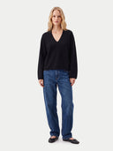 A woman modeling a black long-sleeve V-neck top and blue jeans against a plain light gray studio background.