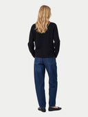 A woman with long blonde hair is shown from behind wearing a black long-sleeve top, blue jeans, and black flats against a light gray studio background.