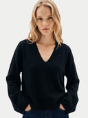 A blonde woman wearing a black long-sleeve V-neck sweater and dark blue jeans, standing against a light gray studio background.