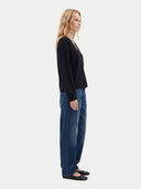 A woman stands in profile wearing a black long-sleeve knit top, blue jeans, and black flats against a plain light gray studio background.