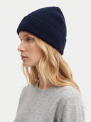 A woman in profile wearing a dark navy ribbed knit beanie and a light gray crewneck sweater, photographed against a clean off-white studio background.