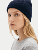 A close-up of a woman wearing a dark navy blue ribbed knit beanie and a light gray crewneck sweater, photographed against a clean, light off-white background.