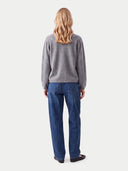 A woman with blonde hair is shown from the back, wearing a gray long-sleeve sweater and blue jeans against a light gray studio background.