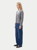 A woman stands in profile wearing a light gray knit cardigan over a white top, blue wide-leg jeans, and black flats, photographed against a plain light gray studio background.