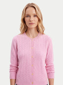 A woman wearing a light pink cable-knit cardigan sweater with gold buttons, photographed against a solid light gray background.